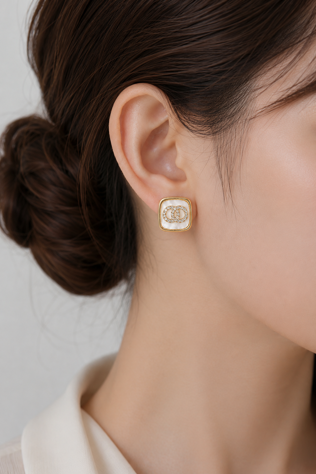 A close-up of a woman's ear wearing a gold square stud earring with a white enamel background and a double-C logo in the center.