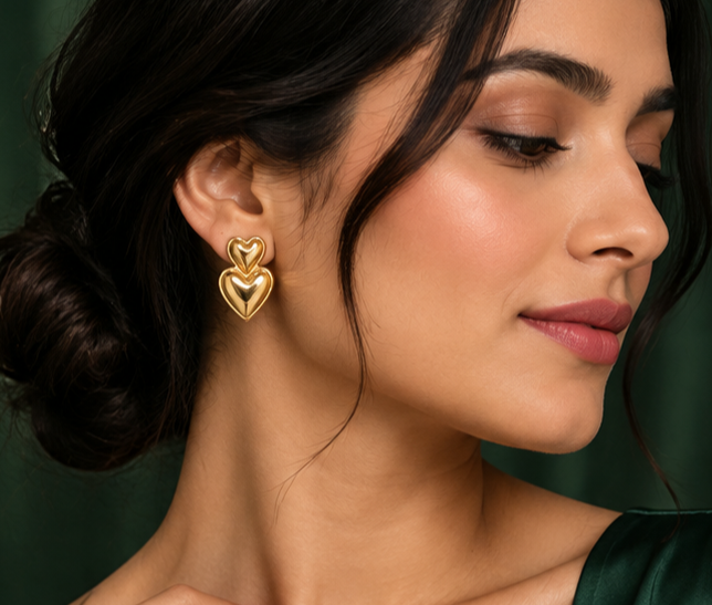 Woman wearing a NAXAYA gold-tone double heart earring, showing the stacked heart design against a dark green background.⠋
