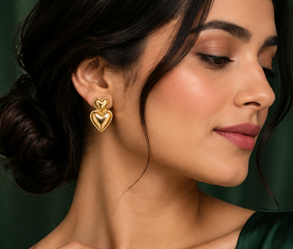 Woman wearing a NAXAYA gold-tone double heart earring, showing the stacked heart design against a dark green background.⠋
