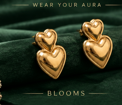 NAXAYA polished gold-tone double heart earrings on dark green fabric with “Wear Your Aura” and “Blooms” text.
