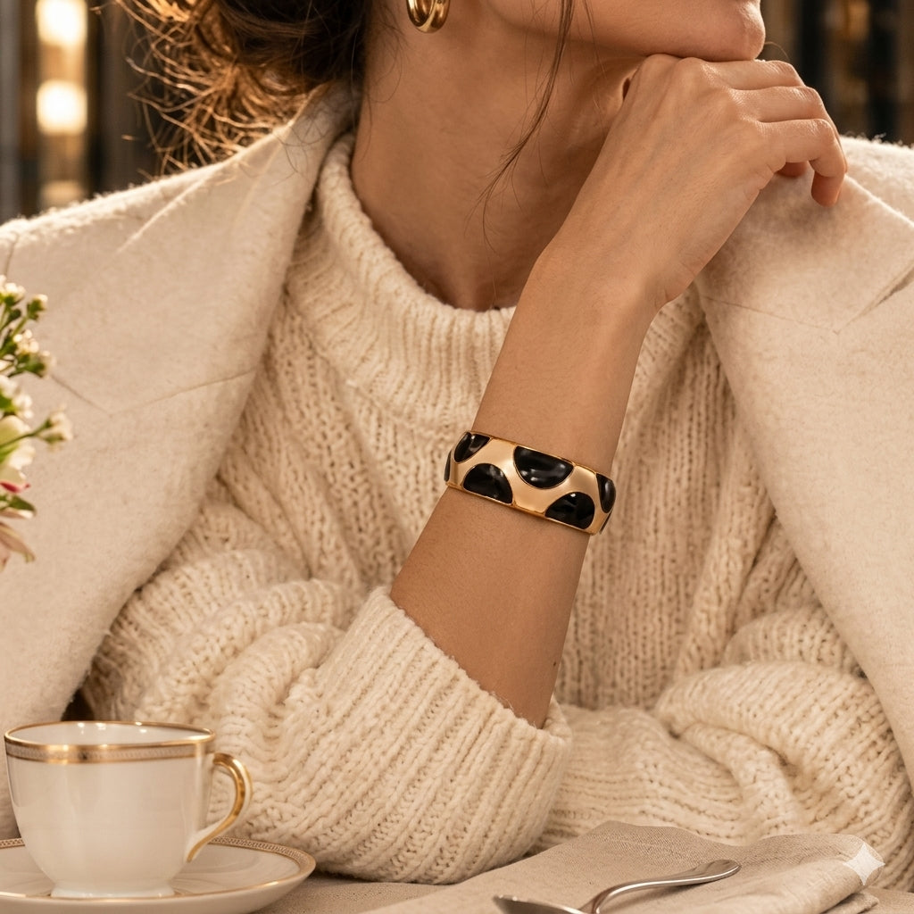 NAXAYA Modern Noir black and gold-tone bracelet styled with a cream sweater and coat at a cafe table.