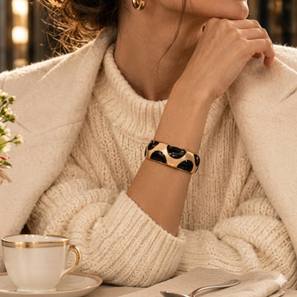 NAXAYA Modern Noir black and gold-tone bracelet styled with a cream sweater and coat at a cafe table.