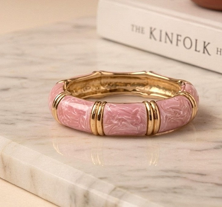 NAXAYA Aura Pink enamel bangle resting on a light stone surface beside a book, showing its gold-tone interior.